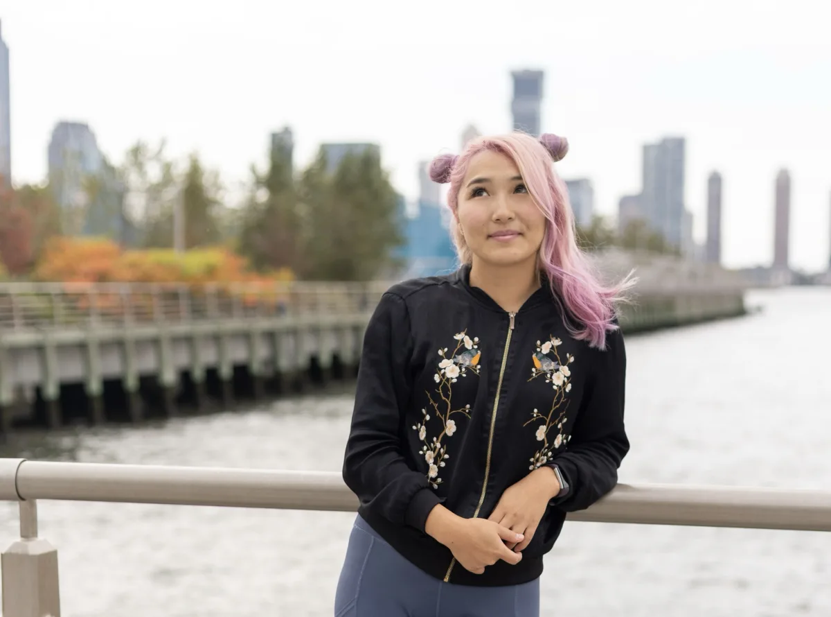 Yanjaa Westgate leaning on a railing by the Hudson River in an embroidered black jacket, with the skyline behind her
