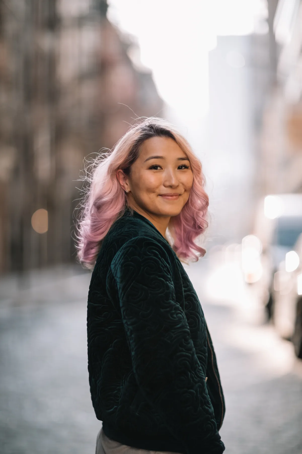 Yanjaa Westgate looking over her shoulder on a cobbled New York street, wearing a dark green velvet jacket, buildings soft in the background