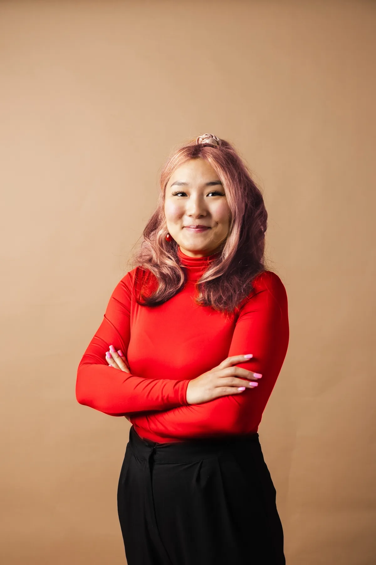 Yanjaa Westgate in a red turtleneck, arms crossed and smiling, against a warm tan studio backdrop