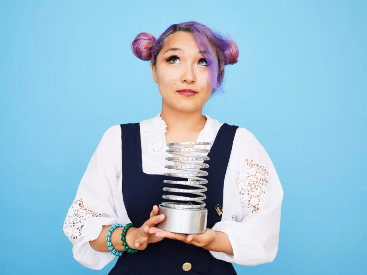 Yanjaa Westgate with purple hair in two buns, holding a silver Webby Award trophy against a sky-blue background